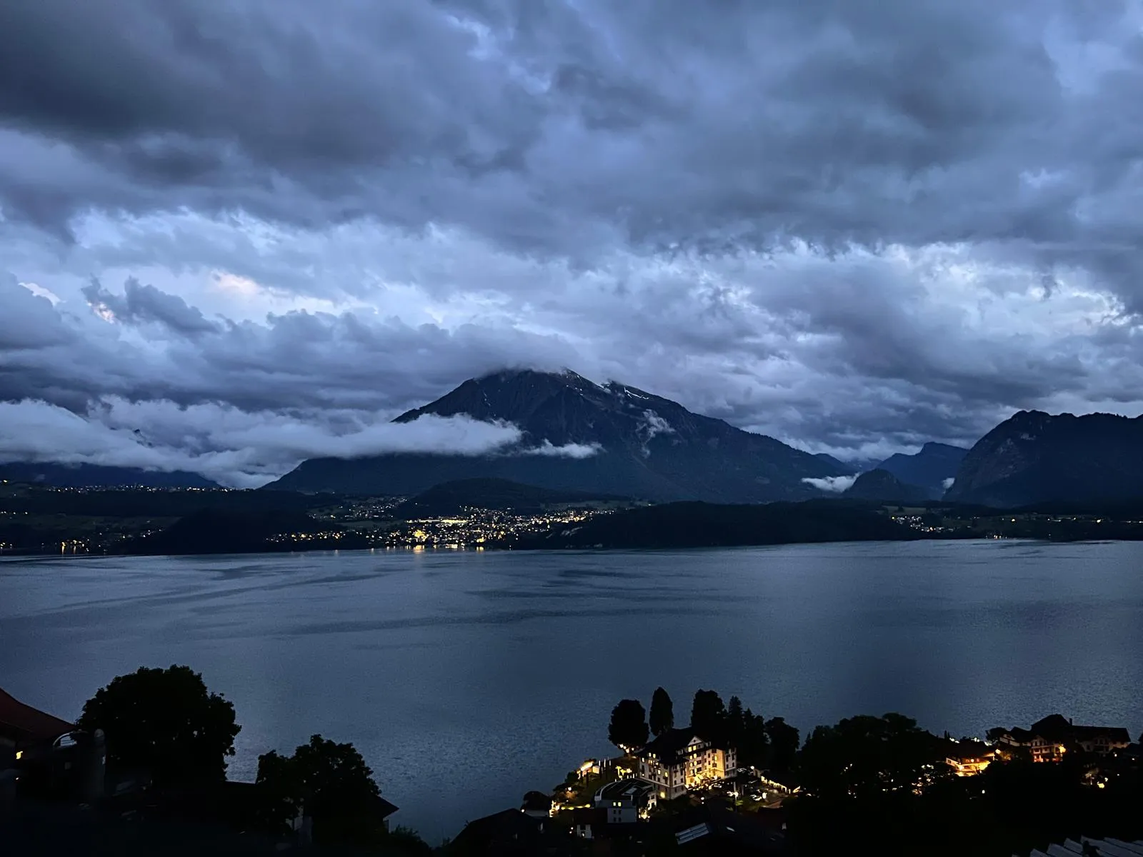 A lake at dusk under a heavy ceiling of cloud, with the lights of settlements along the far shore.