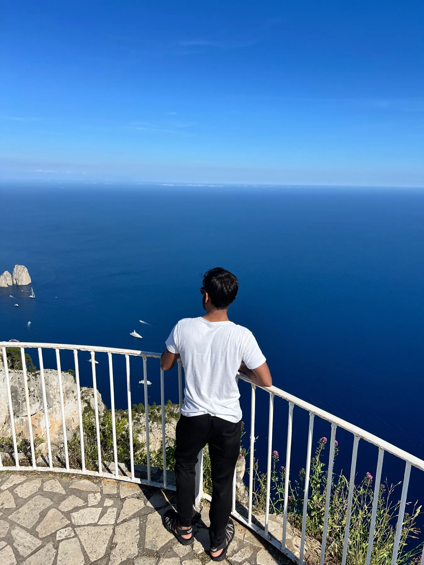 A person seen from behind at a white railing, high above a deep blue sea with small boats and two rock stacks below.